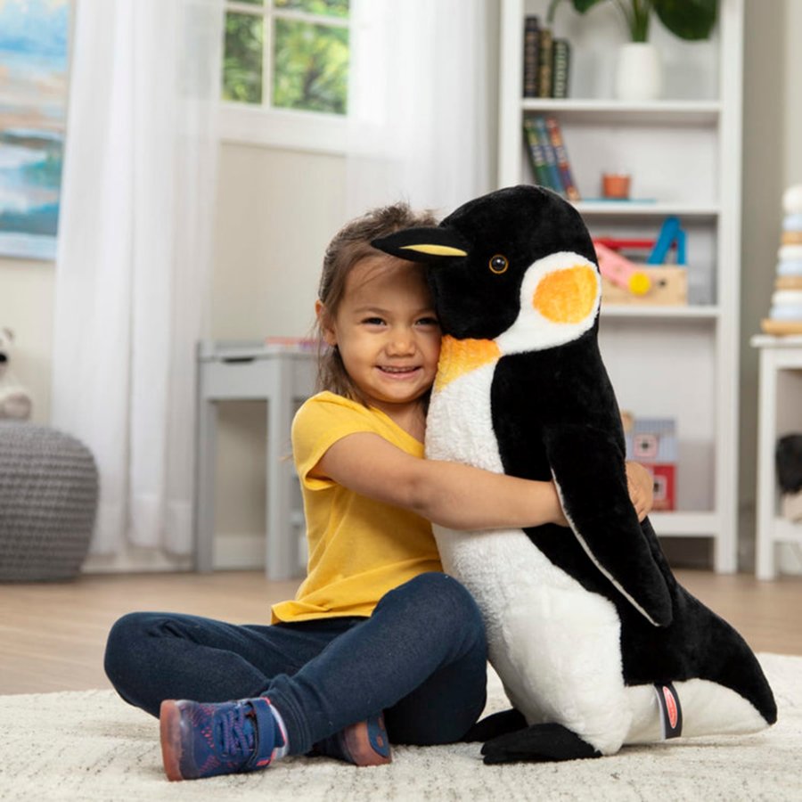 Child holding a large plush penguin toy in a room with books and toys.