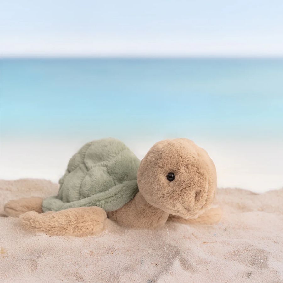 Plush turtle toy on a sandy beach with a clear sky.