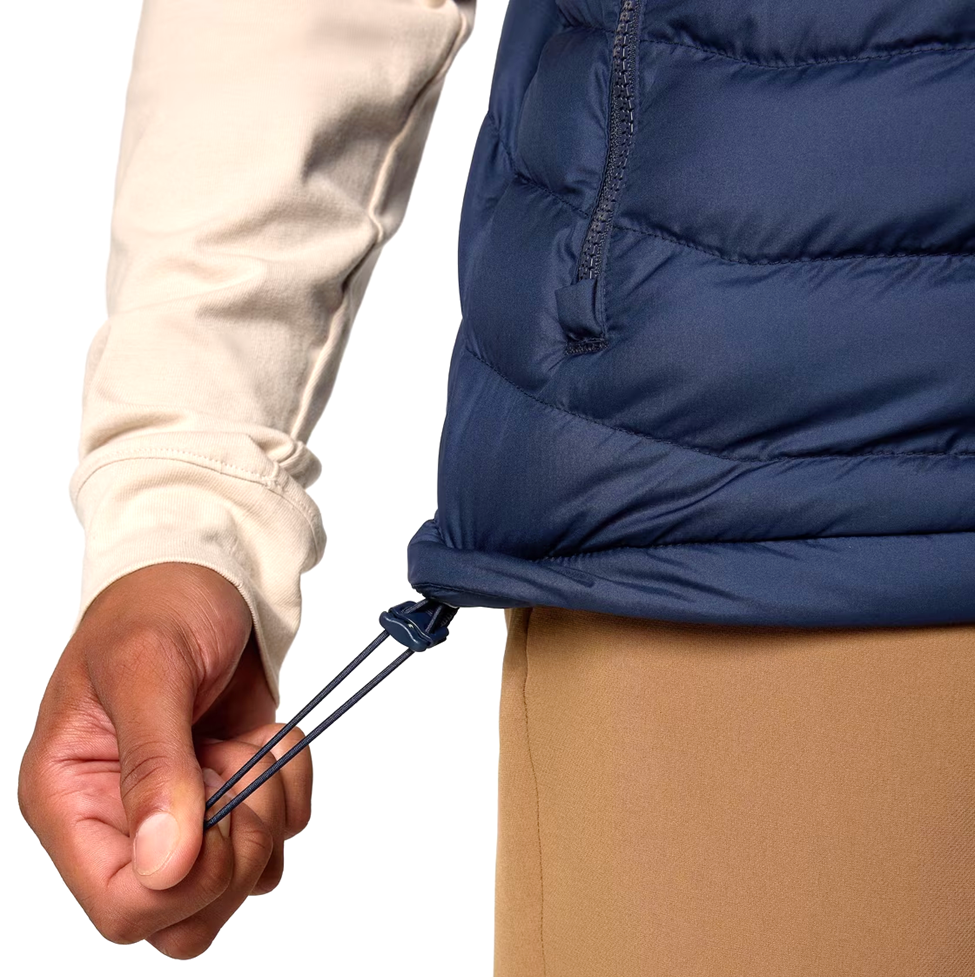 Close-up of a person wearing a blue puffer vest with a beige jacket and brown pants on a white background.