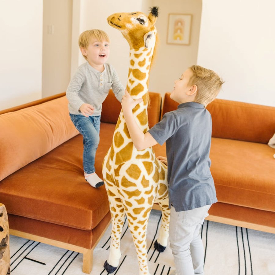 Two children playing with a large stuffed giraffe in a living room.