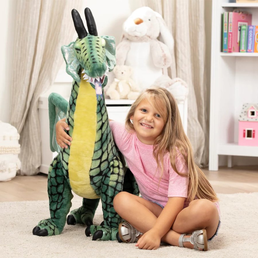 Child holding a large green dragon plush toy in a room with books and stuffed animals.