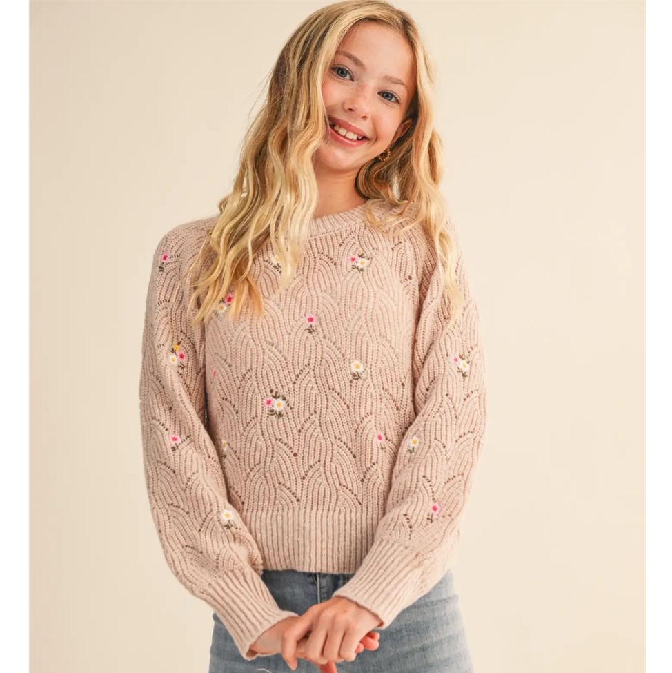 Young girl wearing a beige cable knit sweater with floral embellishments on a beige background