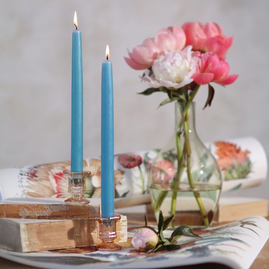 Two blue candles in glass holders on a table with a vase of flowers and a book in the background.