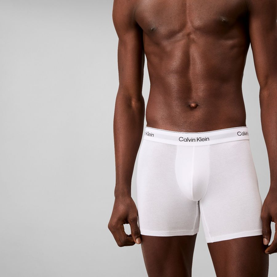 A close-up of a male model wearing white Calvin Klein boxer briefs against a light gray studio background.