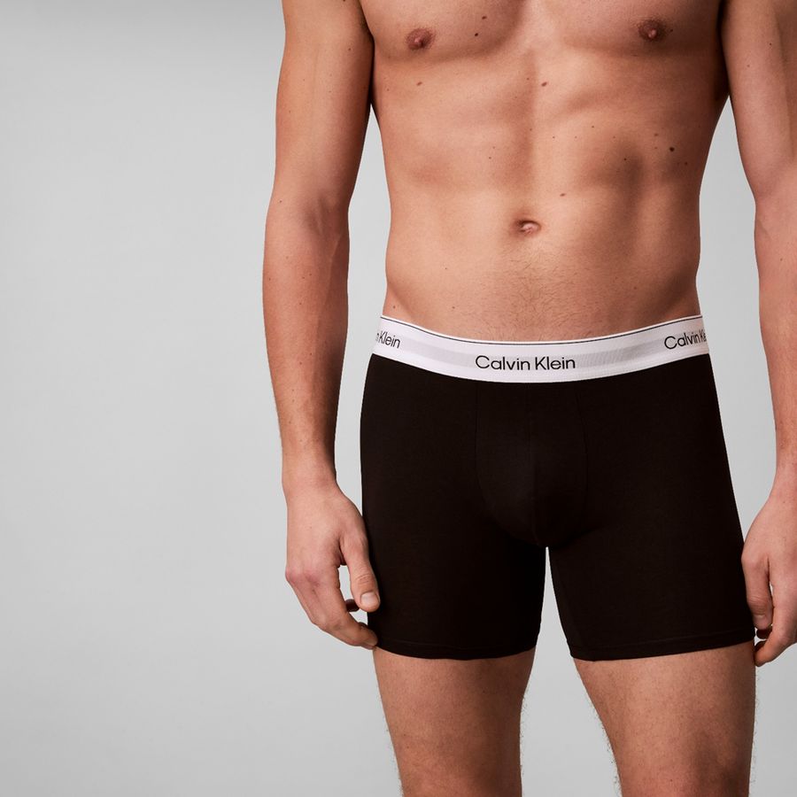 A close-up of a male model wearing black Calvin Klein boxer briefs against a light gray studio background.