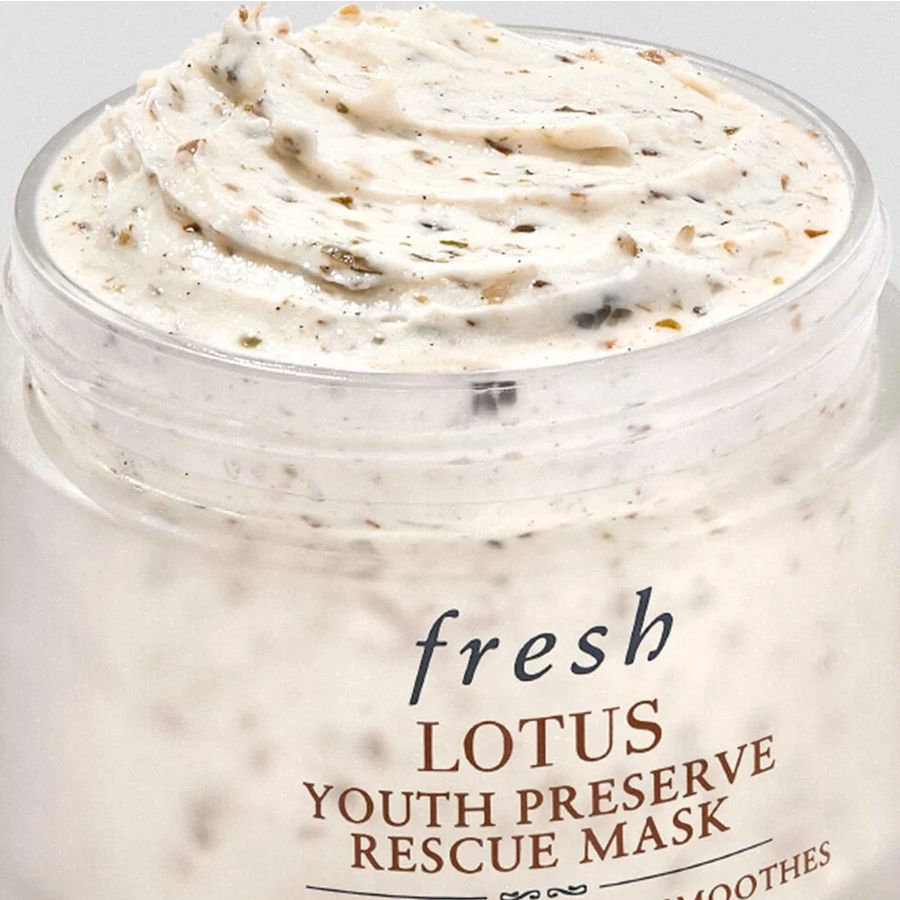 A close-up of a clear jar filled with a thick, swirled off-white cream mask labeled 'fresh LOTUS YOUTH PRESERVE RESCUE MASK'. The mask contains visible small green and brown botanical specks, and the jar is shown against a clean white background.