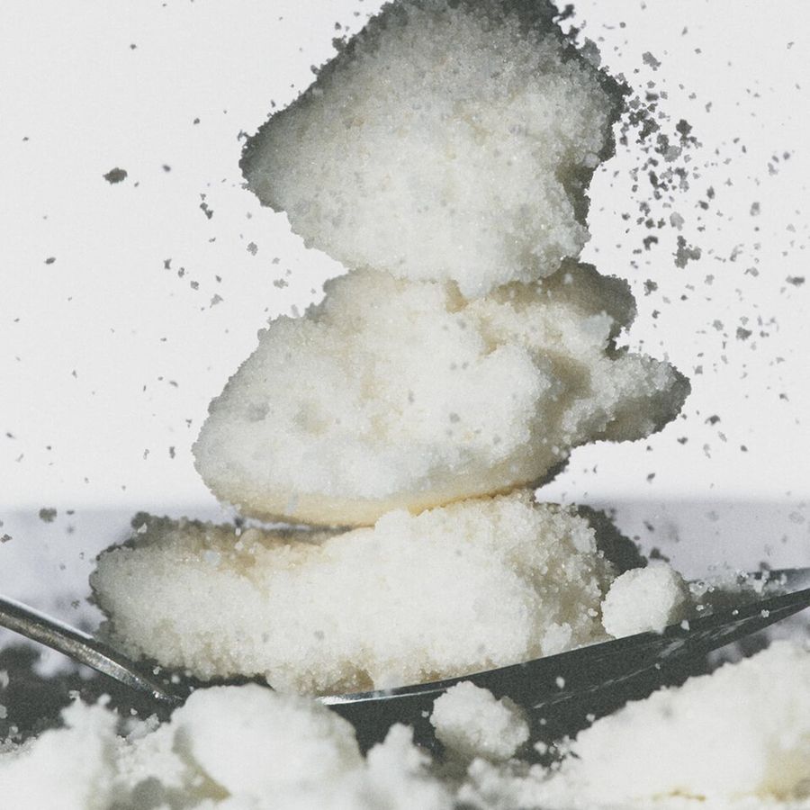A close-up of coarse white sugar crystals stacked on a metal spoon against a white background, with sugar dusting and splashing around the spoon.