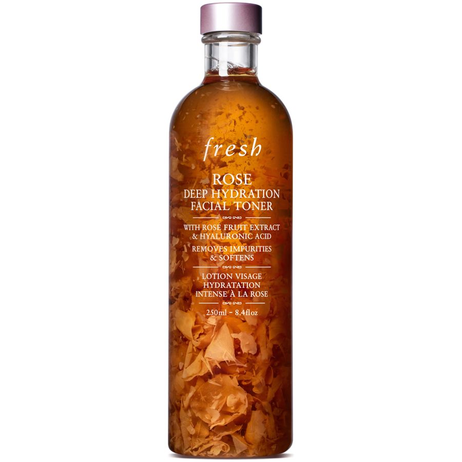 Fresh Rose Deep Hydration Facial Toner in a clear glass bottle with a metallic rose gold cap, filled with amber liquid and rose petals, on a white background.