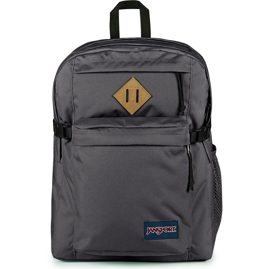 A dark gray JanSport backpack with a tan diamond-shaped logo patch on the front pocket and a blue JanSport brand label near the bottom, shown against a plain white background.