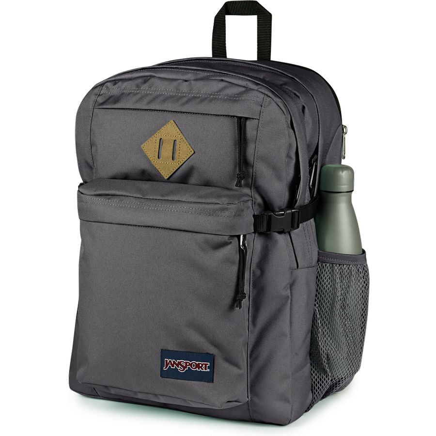 A dark gray JanSport backpack with a front zippered pocket, a diamond-shaped tan patch, a side mesh bottle pocket containing a green bottle, and a JanSport logo patch on the lower front, set against a plain white background.