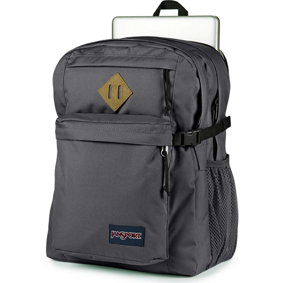 A dark gray JanSport backpack with a gold diamond-shaped patch on the front pocket, a black side strap, and a laptop tucked into the top compartment, shown against a plain white background.