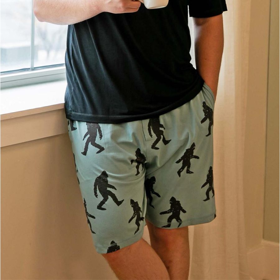 A person wearing sage green shorts with a black 'Bigfoot' footprint pattern, paired with a black t-shirt, standing next to a window.