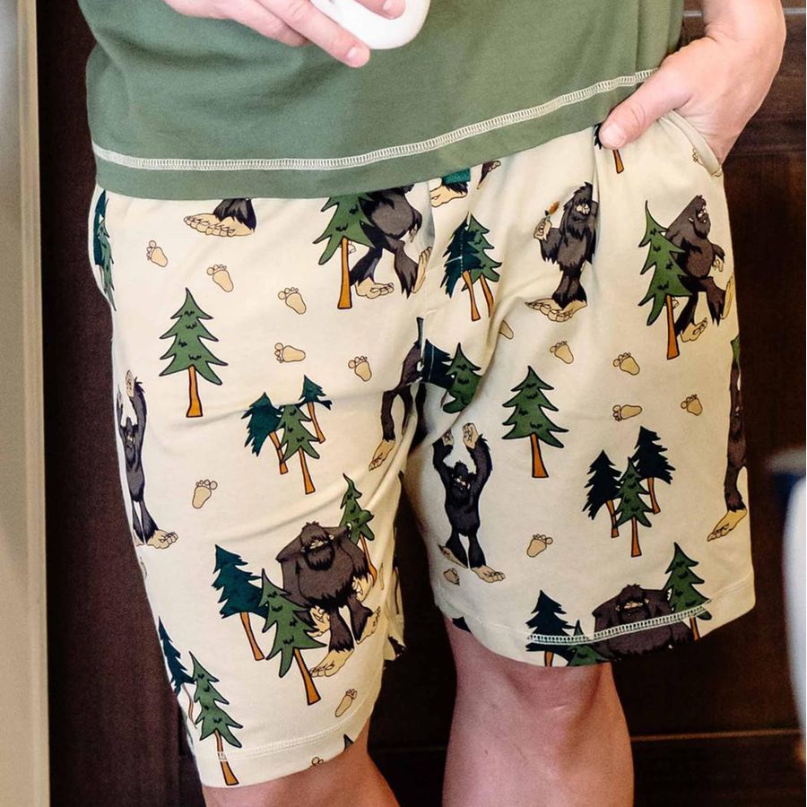 A person wearing beige shorts with a forest and bear print, paired with an olive green shirt. The shorts feature a pattern of pine trees, bears, and footprints on a light beige background.