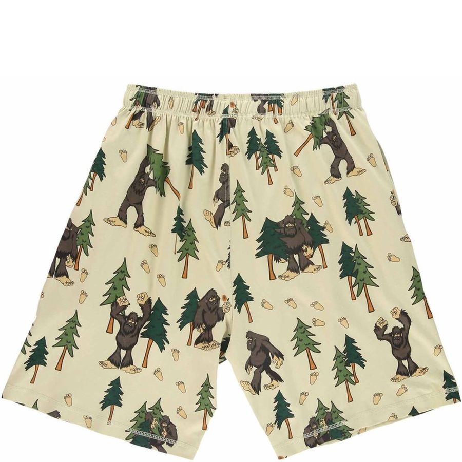 A pair of mens shorts featuring an all-over print of brown Bigfoot figures, green pine trees, and acorns on a light beige background.