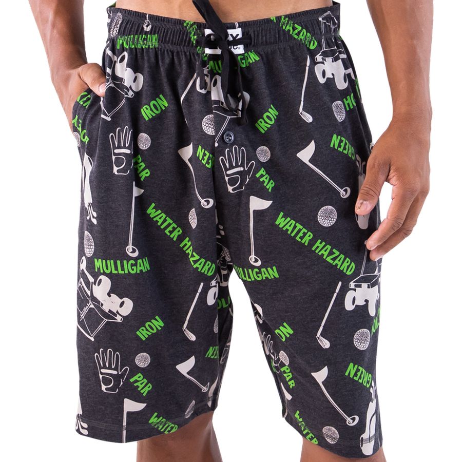 A person wearing dark charcoal heather golf-themed shorts with a green and white print featuring golf clubs, balls, mallets, and text such as 'Mulligan', 'Iron', and 'Water Hazard'.