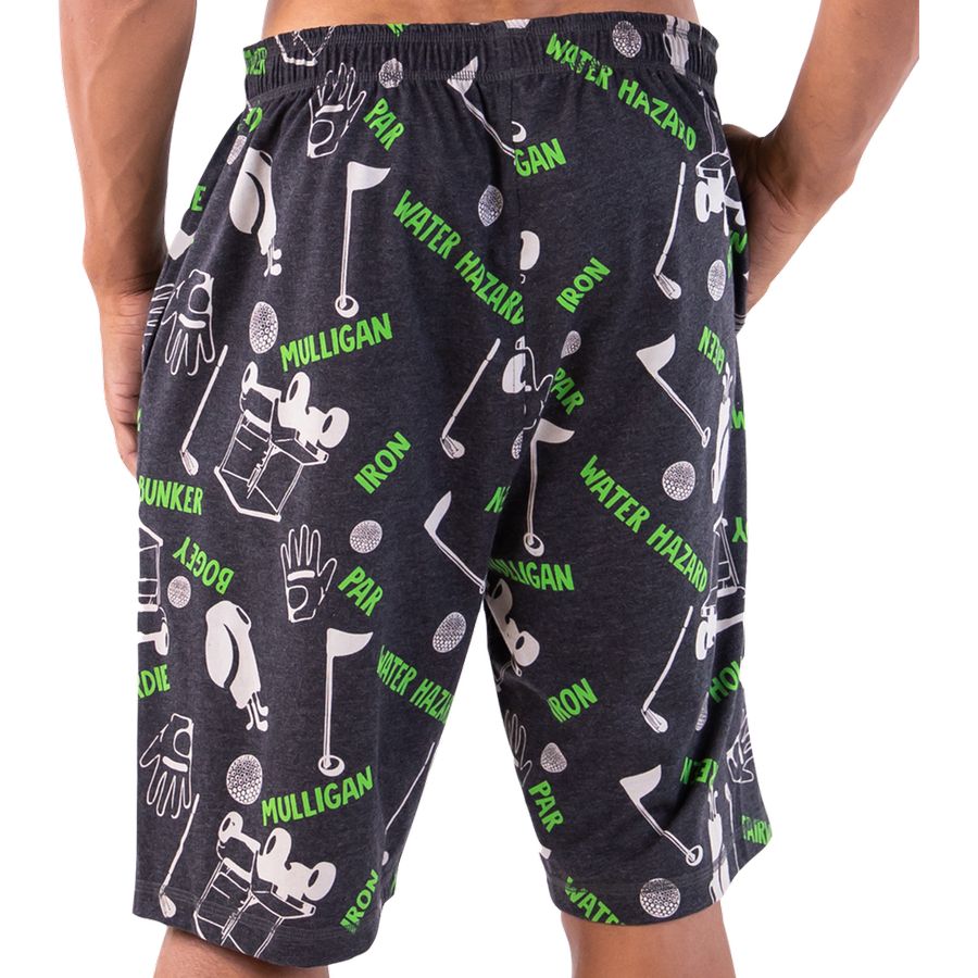 A person wearing dark charcoal heather golf-themed shorts with a green and white print featuring golf clubs, balls, mallets, and text such as 'Mulligan', 'Iron', and 'Water Hazard'.